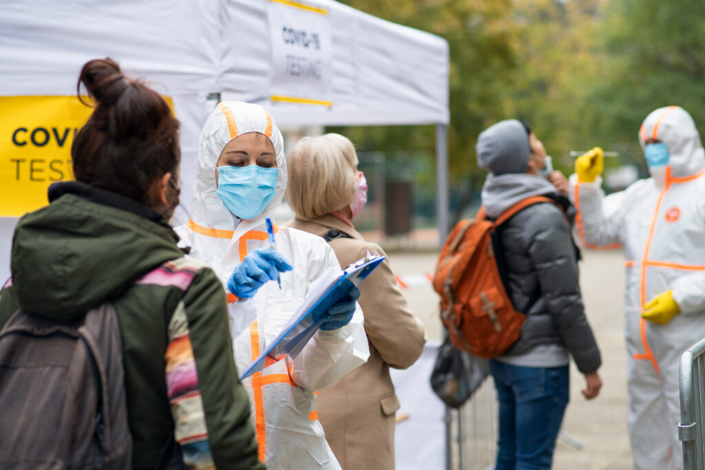 Nurse holding a clipboard while people wait at an outdoor COVID-19 testing center.