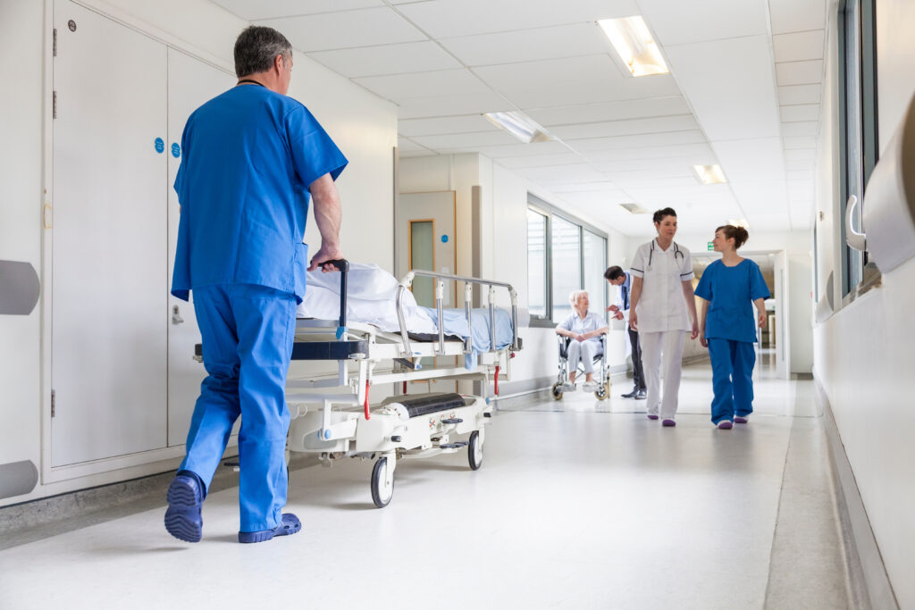 Male nurse pushing a stretcher in a hospital corridor with doctors and nurses talking, and a senior female patient.
