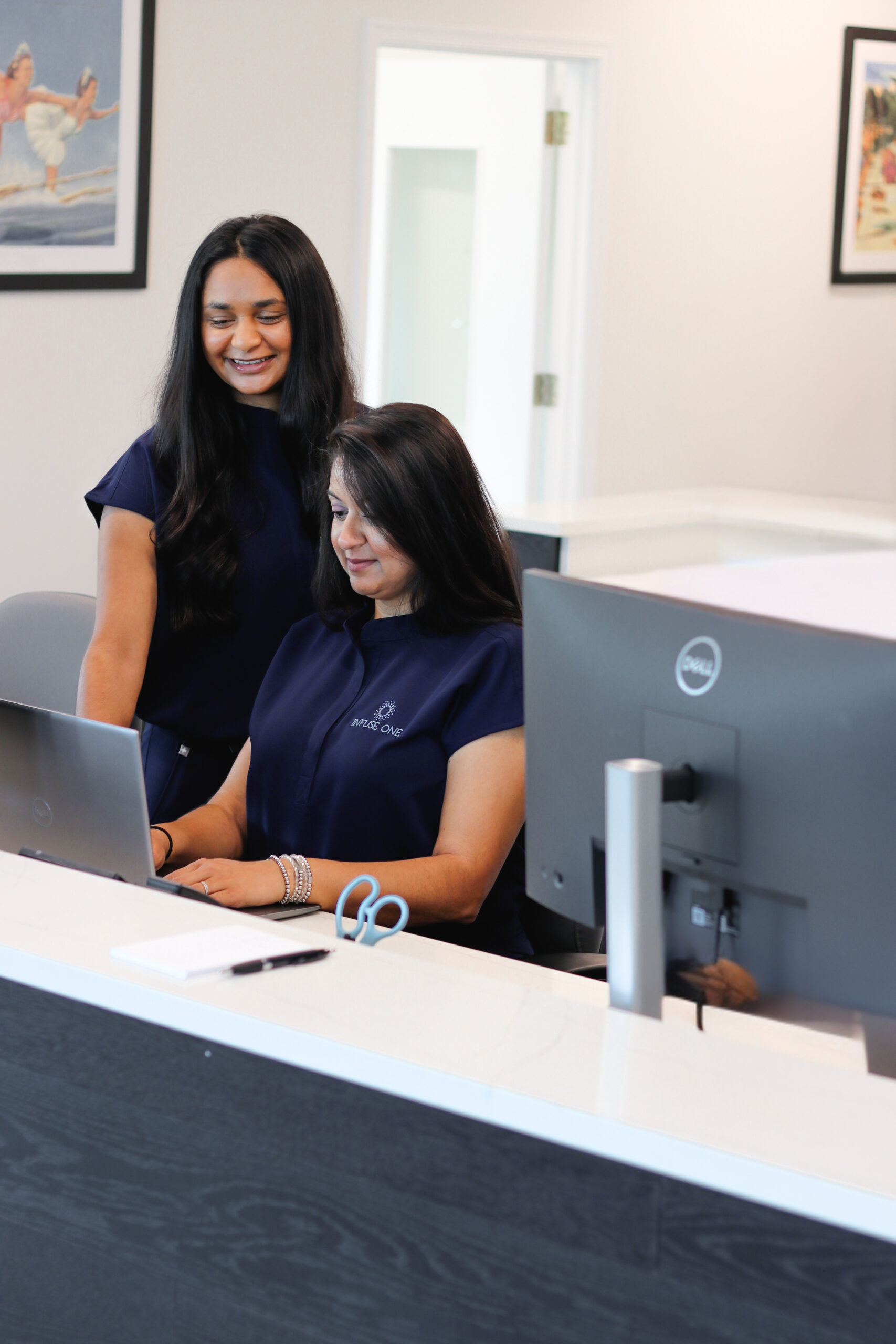 Two women wearing Infuse One shirts huddle around a computer, smiling.