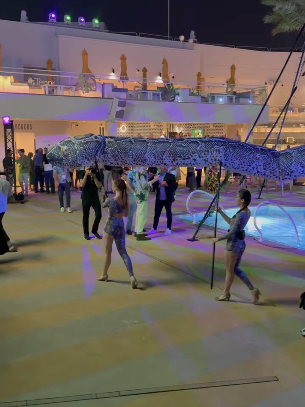 A line of women walk through a party at the pool hoisting a large snake parade puppet.