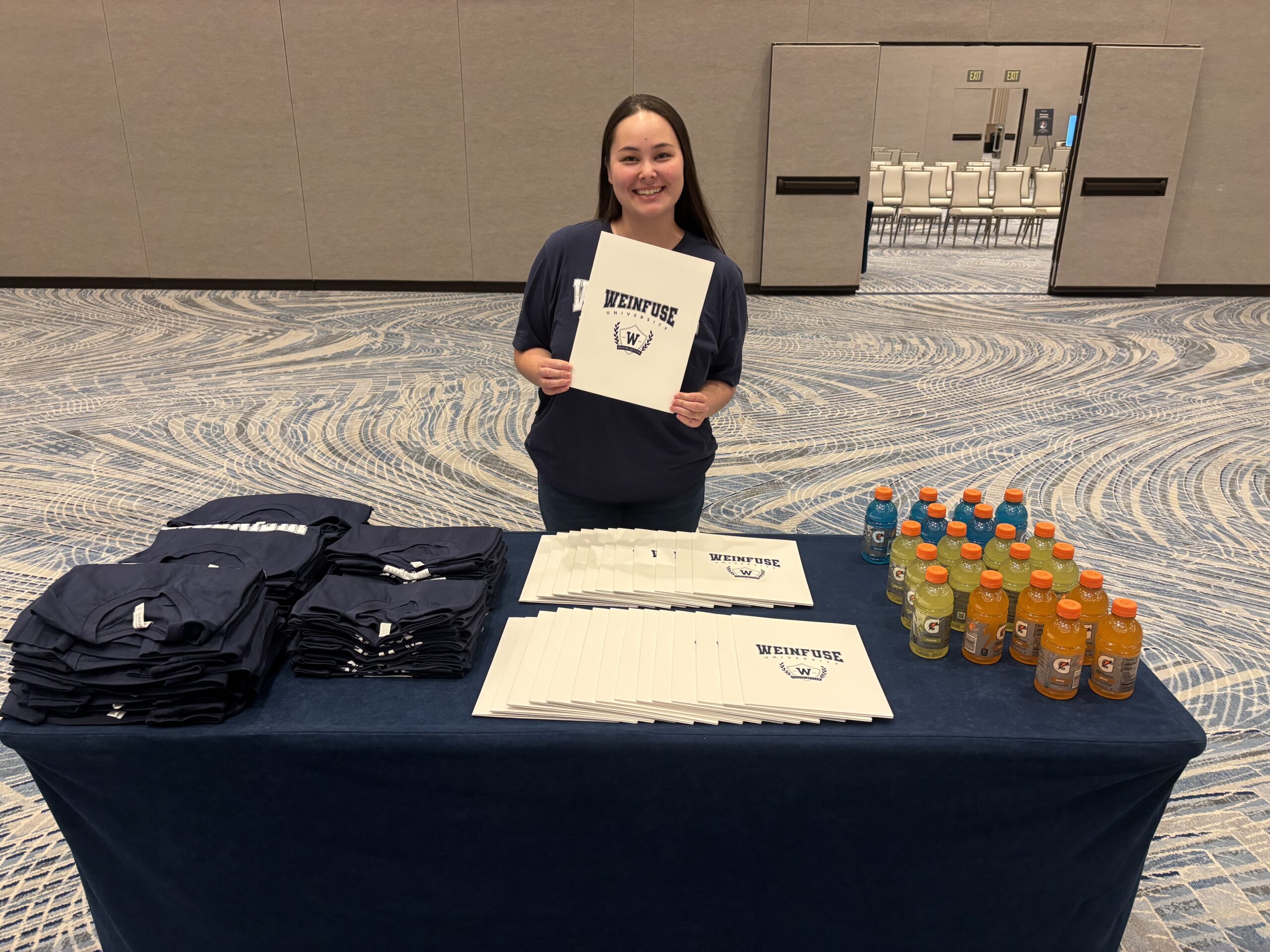 A welcome desk at the 2026 WeInfuse University Users Meeting with t-shirts, folders, and gatorade.