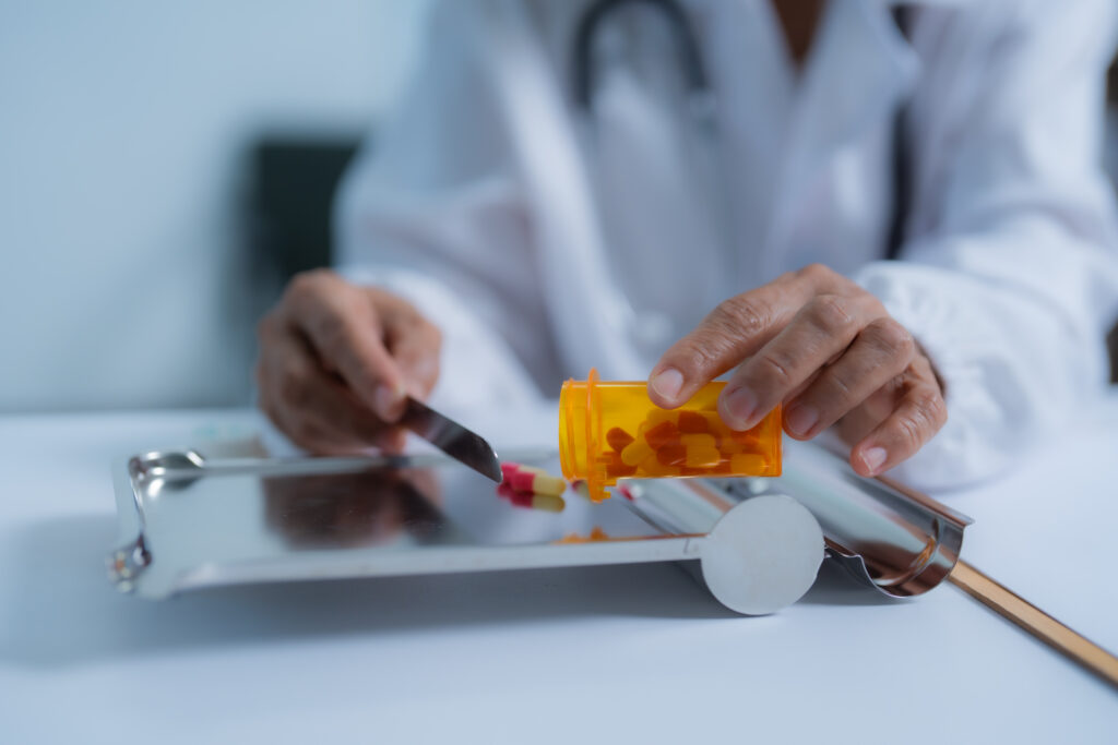 Pharmacy technician wearing lab coat and stethoscope is using a pill counting tray and a metal spatula to prepare medication from a prescription bottle.